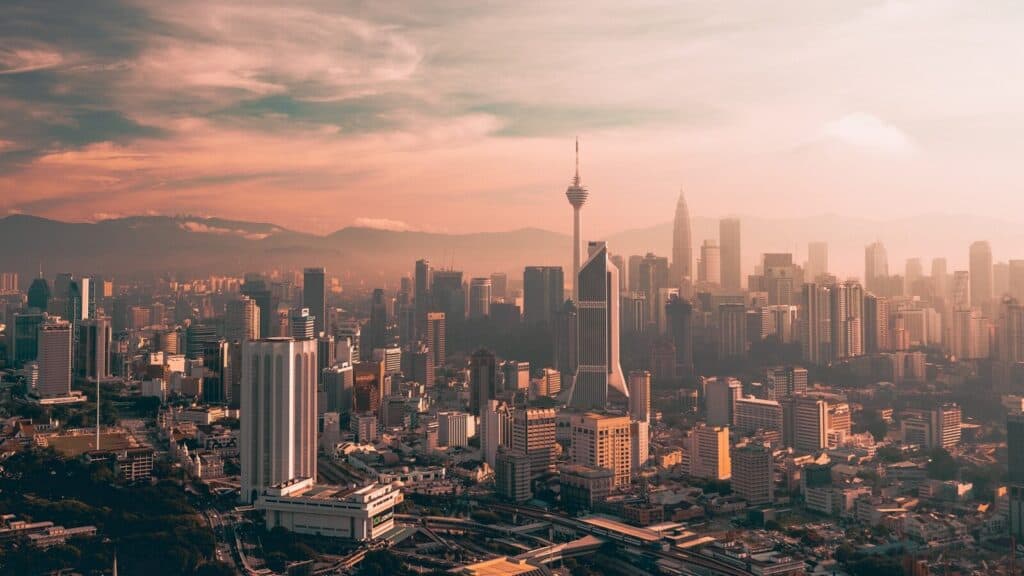 Kuala Lumpur skyline at sunset with skyscrapers, KL Tower, and hazy mountains in the background.
