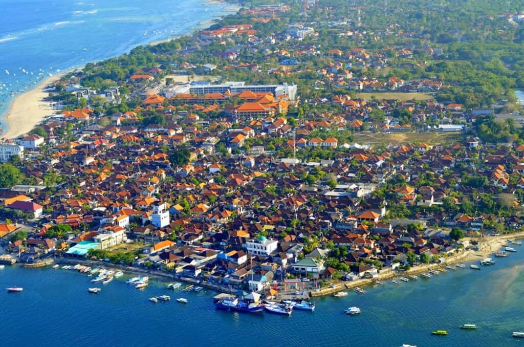Aerial view of a coastal town in Bali with beaches, boats, red rooftops, and tropical greenery.