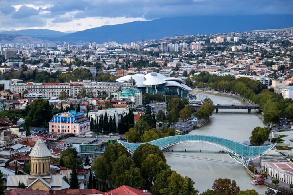 View over Tbilisi with the Kura River, modern bridge, city buildings, and mountains in the background.