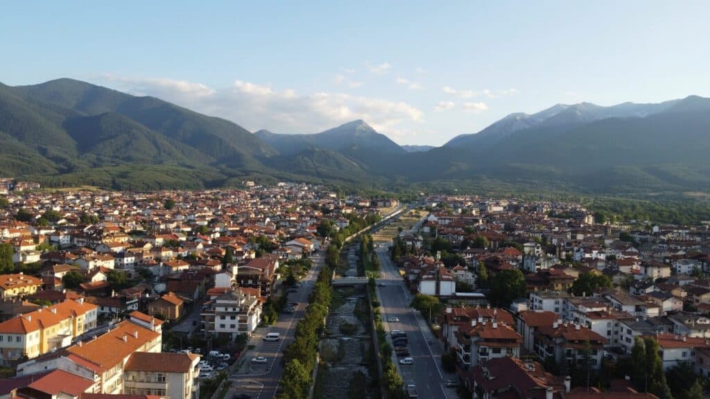 View over Bansko with red-roofed buildings, a long road, and mountains in the background.