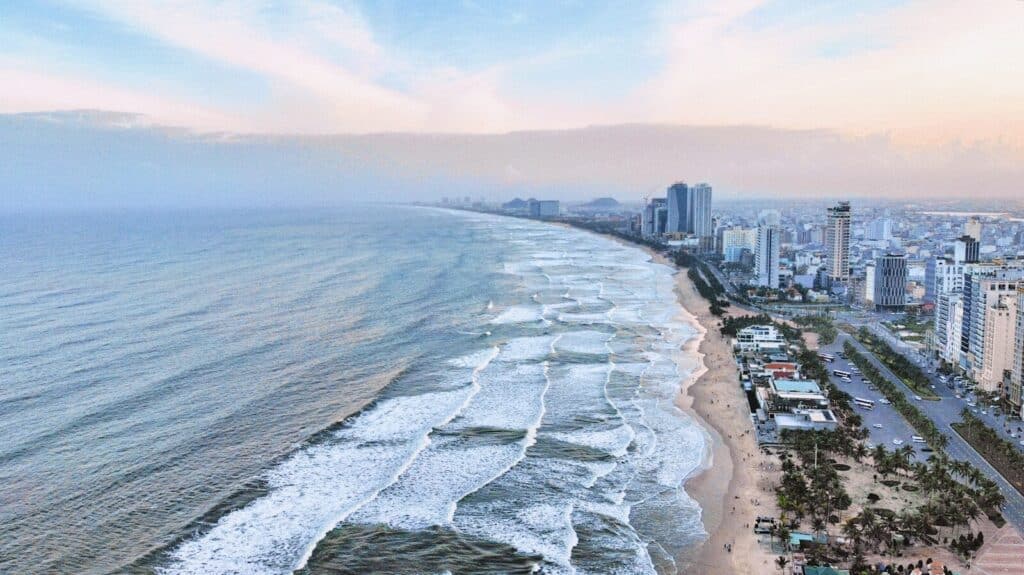 Aerial view of Da Nang coastline with beach, waves, high-rise buildings, and a coastal road.