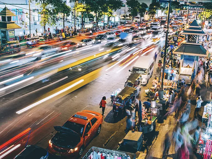 Busy Thai traffic at night with cars, scooters, street vendors, and pedestrians.